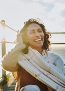 Mature woman wearing a pashmina laughing while sitting at a scenic viewpoint by the ocean at sunset