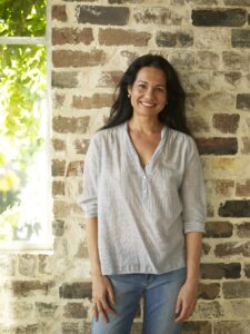 A standing smiling Tess in a green shirt front of a brick wall with a window and a tree outside