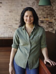 A standing smiling Tess in a green shirt front of a brick and wooden floor background
