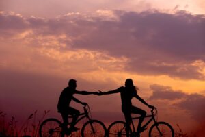 Silhouette of a man and women riding bikes holding hands in front of a beautiful sunset